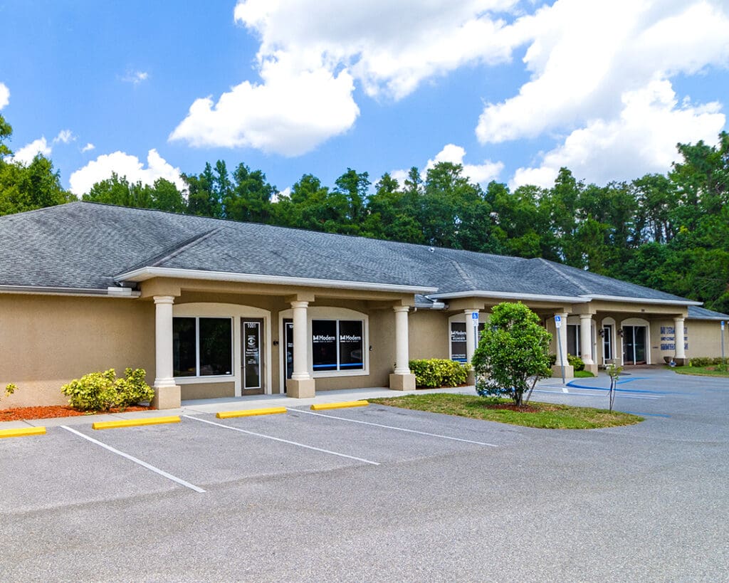 Podiatrist Tampa's Lutz branch offers a contemporary beige building facade. The large windows display business hours and contact information. The entrance is flanked by decorative columns supporting a protruding roof, and the ground near the doorway is adorned with potted plants.