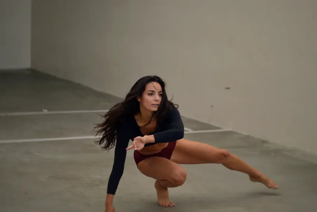 Woman in black long sleeve shirt and bikini bottom crouched down in a dance move with one leg out inside an industrial space with concrete floor