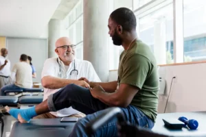 White male doctor with stethoscope seated shakes hand with black adult male on patient table in green shirt and jeans and foot wrapped for podiatric treatment
