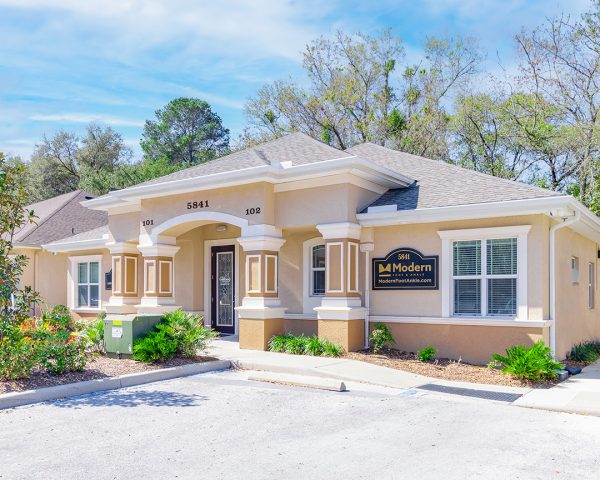 Podiatrist Tampa office in Wesley Chapel, showcasing a single-story beige structure, twin front entrances with the numbers 101 and 109 prominently displayed, a symmetrical design with columned porticos, and a neatly organized parking area set against a backdrop of tall trees.