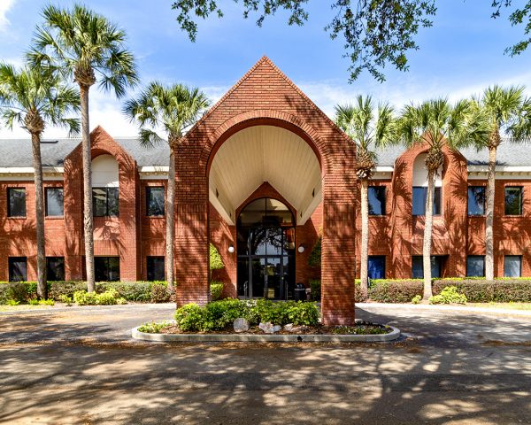 Podiatrist Orlando office in Lake Mary with a brick façade, arched entryway, a white sloping roof, tall palms flanking the building, and a blue USPS mailbox at the front.