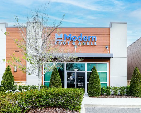 Modern Foot & Ankle podiatrist office in Winter Garden Orlando featuring a bright orange facade with the clinic's logo, clear glass entrance, and young tree on the side.